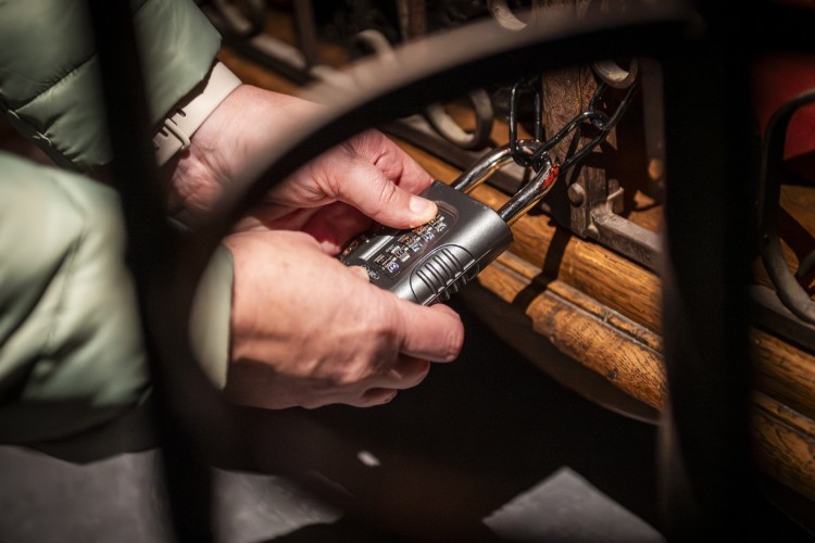 Een hand houdt een cijferslot vast terwijl het wordt afgesloten aan een metalen structuur. De achtergrond toont een houten oppervlak en andere elementen die deel uitmaken van de omgeving.