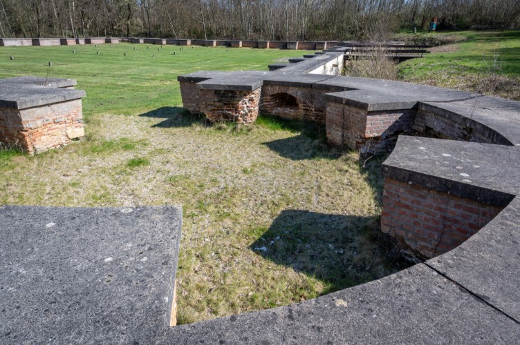 Een uitzicht op een deel van een historische ruïne met bakstenen muren en betonnen elementen, omringd door gras en bomen. De ruimte lijkt te zijn ingericht voor herdenking of educatieve doeleinden.