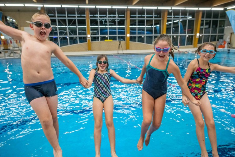 Vier kinderen springen samen in een zwembad, met zwembrillen op en vrolijke gezichten. Ze houden elkaars handen vast terwijl ze in de lucht hangen boven het water.