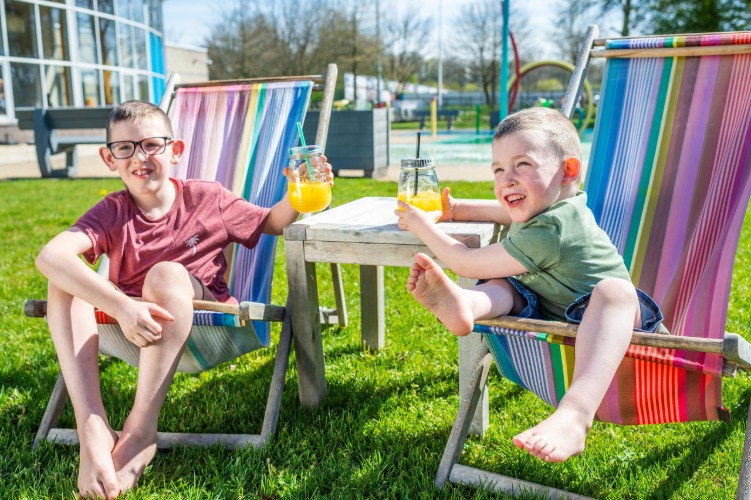Twee kinderen zitten op kleurrijke ligstoelen in een groene tuin. Ze genieten van een zonnige dag en houden glazen met sinaasappelsap vast. Achter hen is een zwembad zichtbaar, omringd door bomen en speeltoestellen.