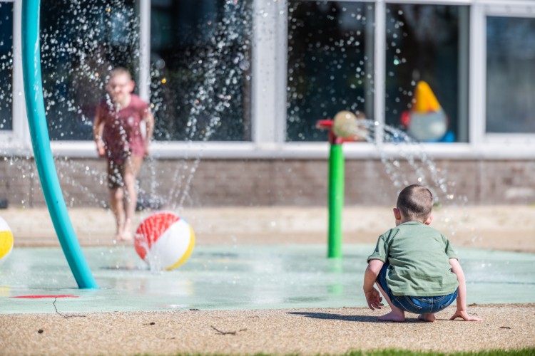Een jong kind zit op de grond in een waterpark, terwijl een ander kind op de achtergrond speelt. Er zijn kleurrijke strandballen en waterstralen die omhoog spuiten. De omgeving is zonnig en vrolijk.