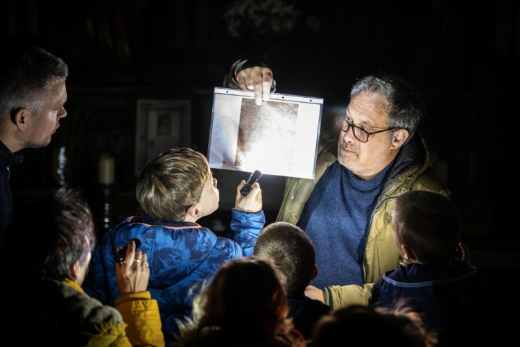 Een man toont een afbeelding aan een groep kinderen, terwijl hij een zaklamp gebruikt om het beeld beter zichtbaar te maken. De setting is donker, wat de aandacht op de afbeelding en de interactie tussen de man en de kinderen vestigt.