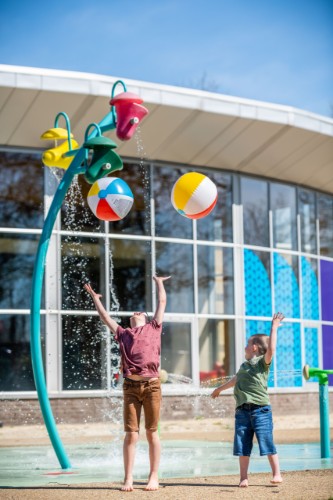 Twee kinderen spelen in een spetterend waterpark. Eén kind staat met zijn armen omhoog onder een waterspeeltoestel, terwijl het andere kind naast hem vrolijk kijkt. Op de achtergrond zijn kleurrijke ballen te zien die door de lucht vliegen en een moderne gebouwstructuur. De zon schijnt helder op deze zomerse dag.