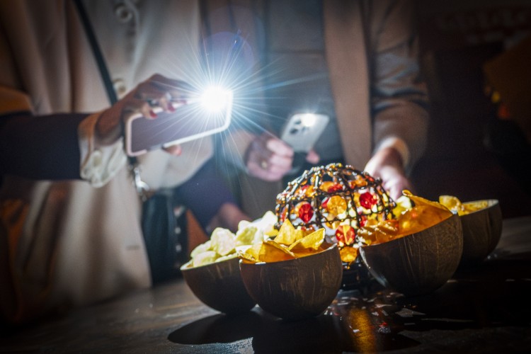 Een hand houdt een smartphone met flits boven een tafel met verschillende kommen chips en een decoratieve lamp in het midden. De omgeving is donker, waardoor de lichtstralen van de flits opvallen.